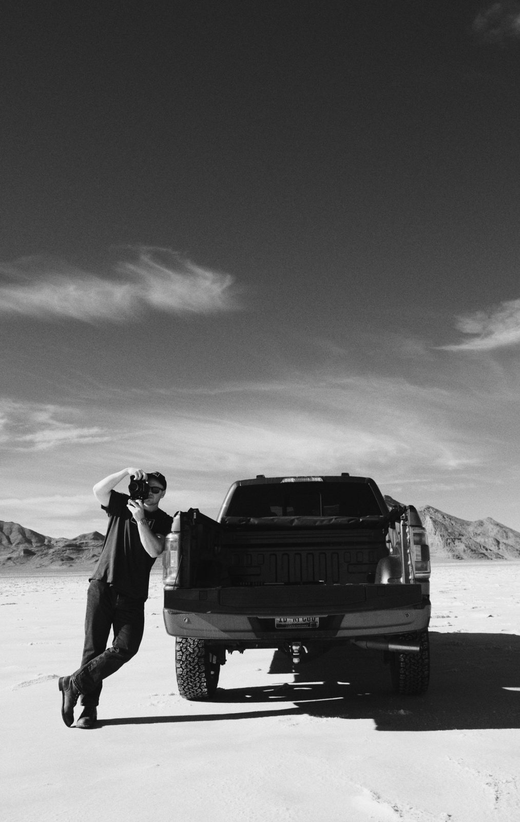 Black-and-white photograph of Clay leaning on the tailgate of a pickup truck on a desert flat, raising a camera, with mountains behind