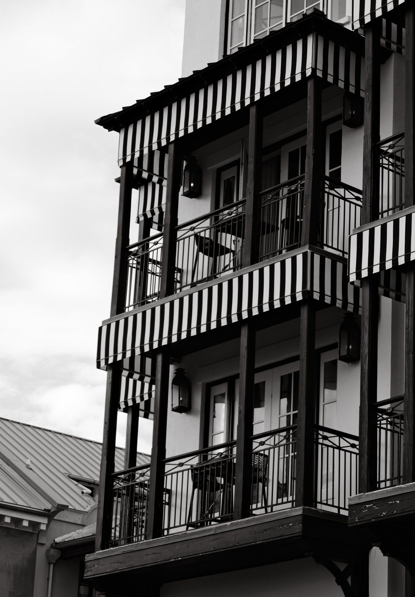 Black-and-white photograph of a hotel facade with stacked balconies, striped awnings, wrought-iron railings and lanterns against a pale sky