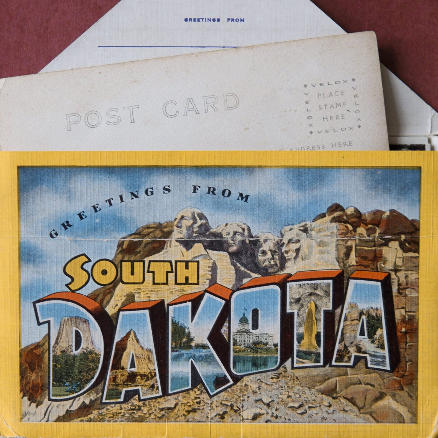 Vintage Greetings from South Dakota postcard folder with blank postcards tucked behind it, on a deep red surface